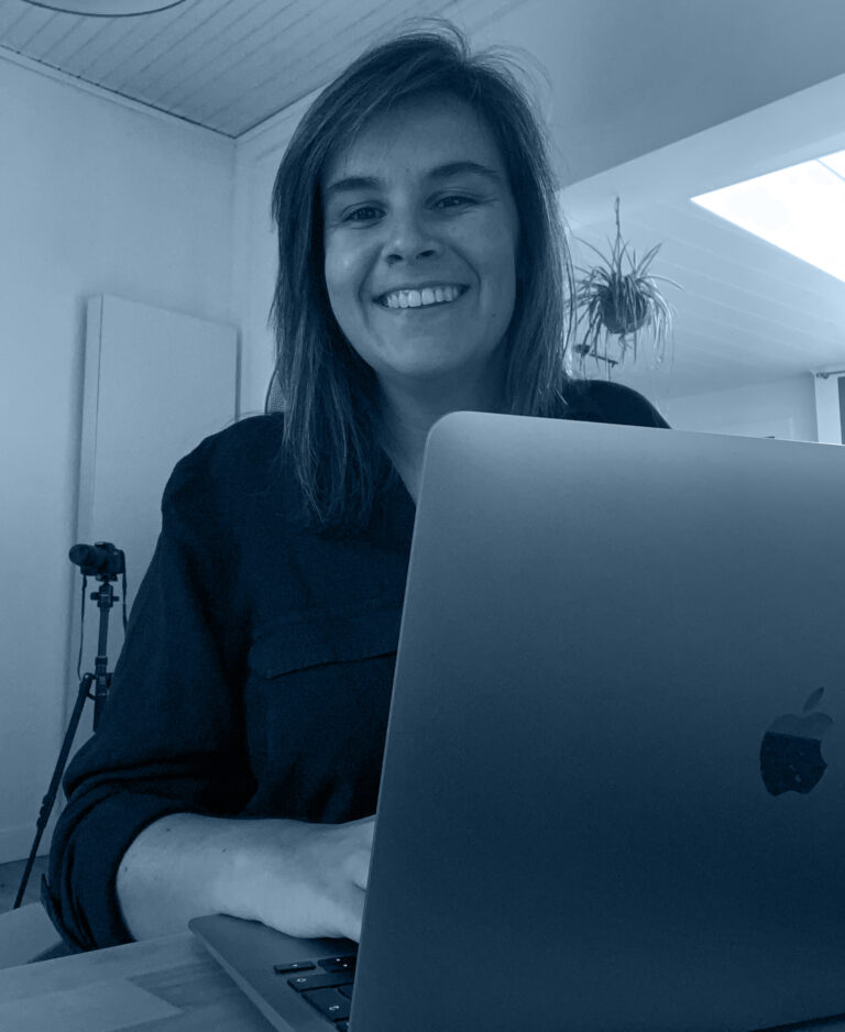 Zelfportret van Eleni achter een laptop lachend met op de achtergrond een fototoestel op een statief.
